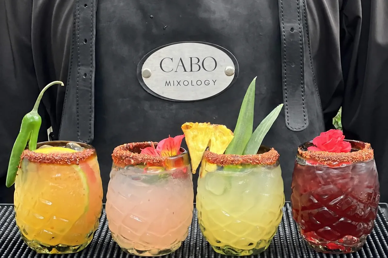 Four colorful cocktails with citrus, chili rims and floral garnishes in front of a Cabo Mixology apron