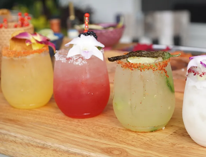 Colorful cocktails with flowers, citrus and chili rims on a wooden bar.