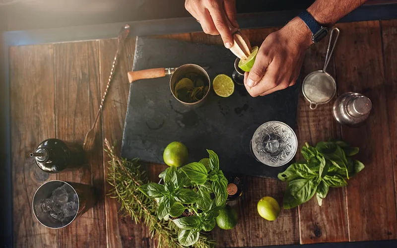 Fresh lime being squeezed beside herbs, cocktail tools and glasses
