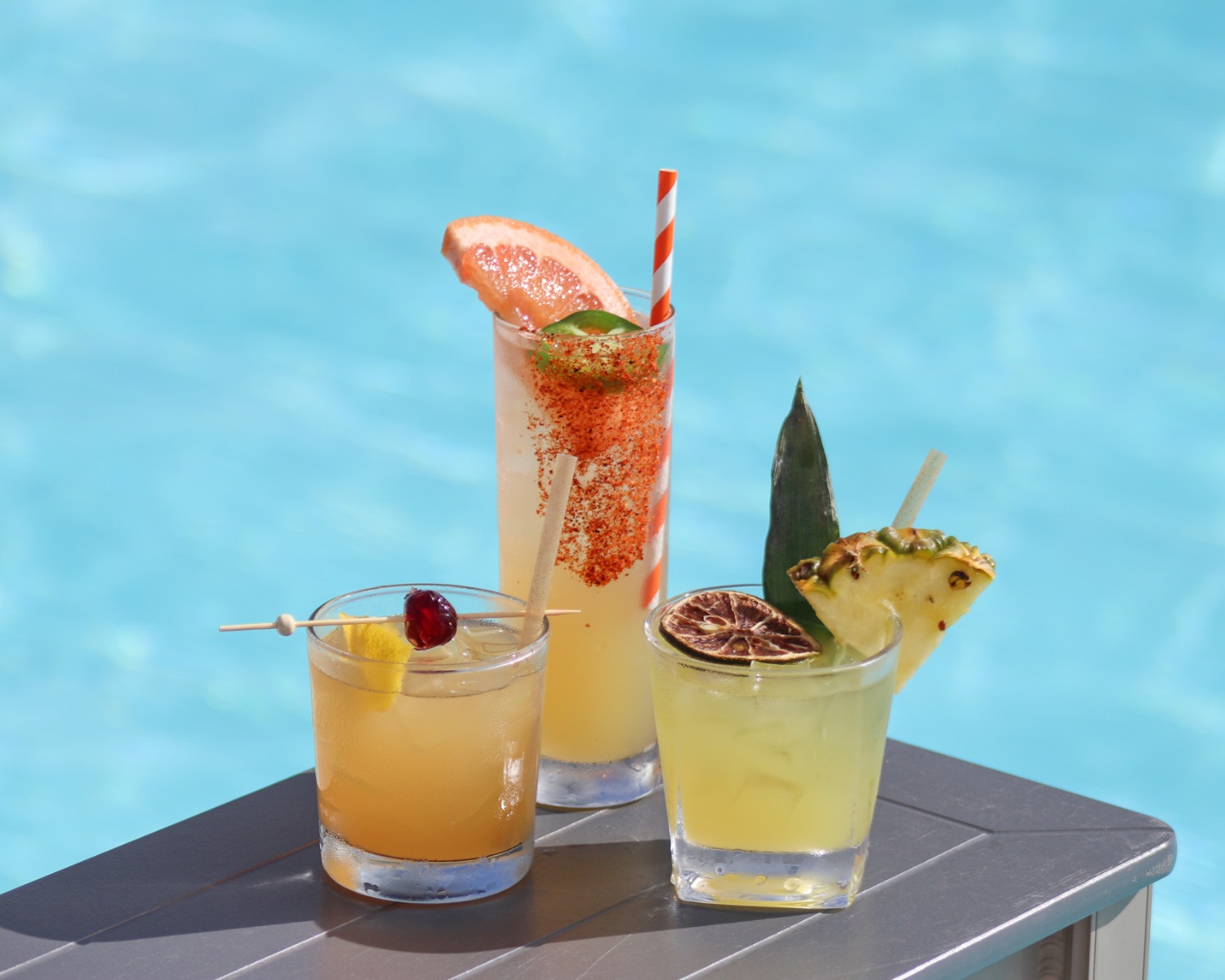 Three citrus cocktails beside a bright blue villa pool